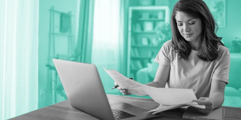 A woman wearing a blue shirt sits at a wooden table in her living room, reviewing documents next to an open laptop.
